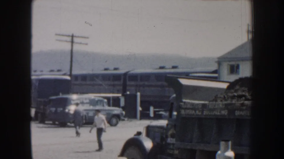 1959 Nebraska home-movie frame with a man crossing a rail-side yard near vintage cars, a train, and a loaded truck.