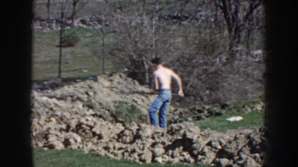 1959 Nebraska home-movie frame showing a shirtless man in jeans digging outdoors near rocks, grass, and leafless trees.