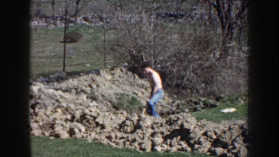 1959 Nebraska 8mm frame of a shirtless man bending to dig in a grassy, rocky yard with brush and bare trees behind him.