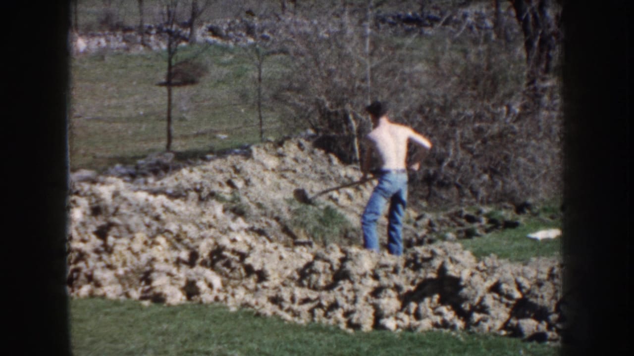 In 1959 Nebraska home-movie footage, a shirtless man in jeans works with a shovel beside a dirt mound in a rural yard.