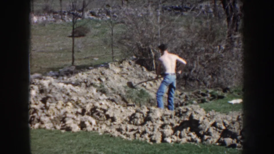 In 1959 Nebraska home-movie footage, a shirtless man in jeans works with a shovel beside a dirt mound in a rural yard.