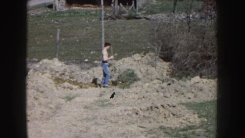 The man stands on or near the dirt pile with a shovel, framed by grass, brush, and fence posts in rural Nebraska footage from 1959.