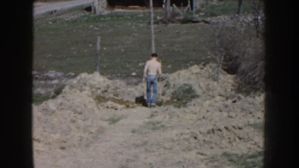 Seen from behind, the shirtless man stands on the dirt mound in a grassy Nebraska yard, with brush and fence posts beyond him.
