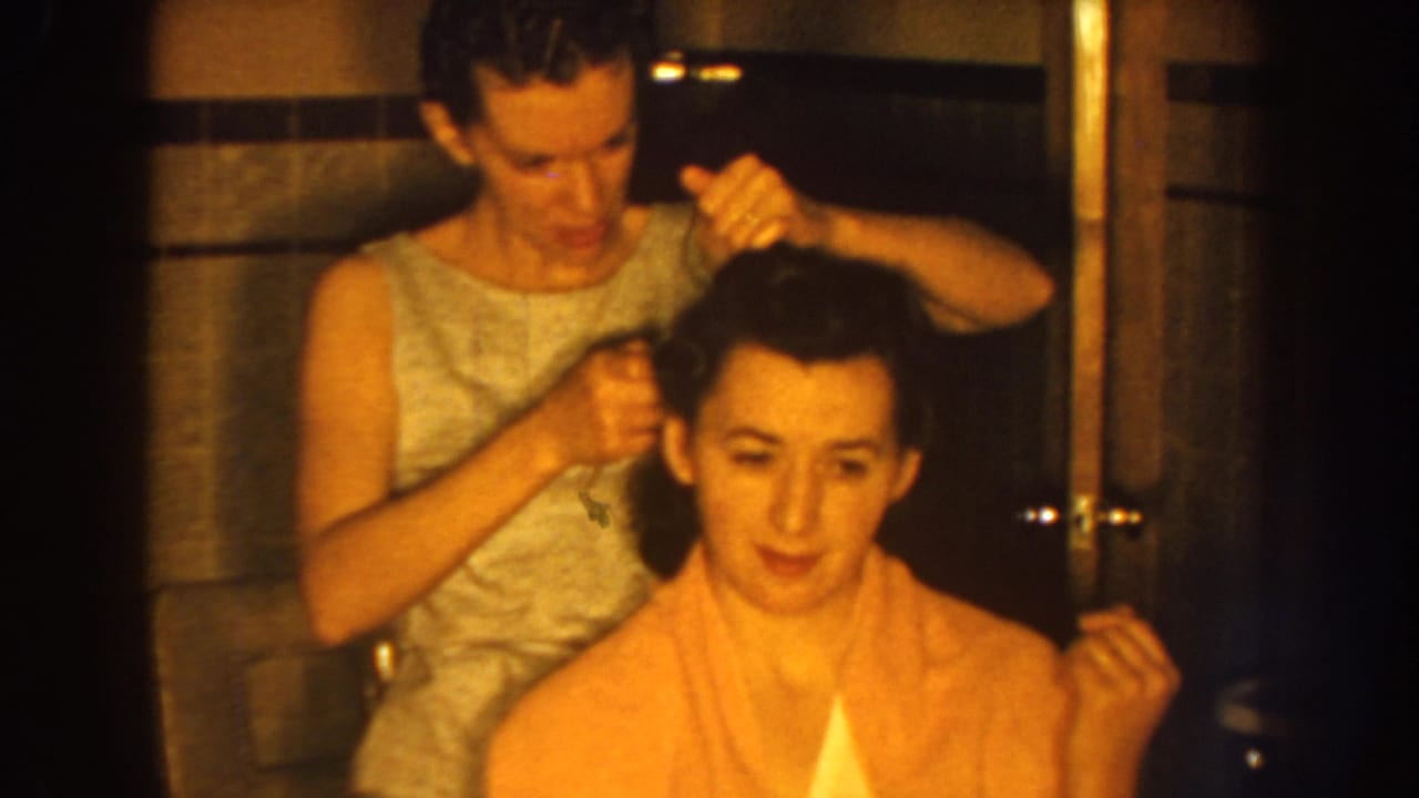 In 1959 Nebraska home-movie footage, a woman in a light sleeveless dress combs the dark hair of a seated woman wrapped in a pink towel.