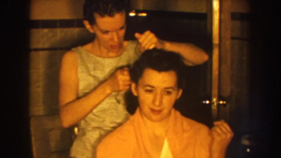 In 1959 Nebraska home-movie footage, a woman in a light sleeveless dress combs the dark hair of a seated woman wrapped in a pink towel.
