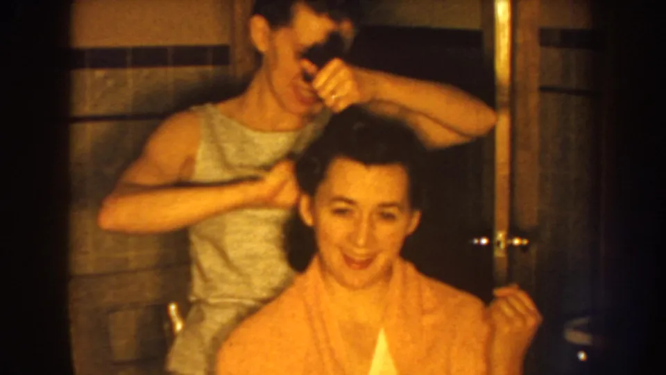 A seated woman smiles during the grooming moment while another woman combs her hair in the warm-lit 1959 Nebraska bathroom scene.