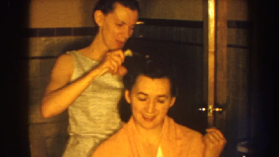 Both women smile during the informal hair-combing scene, with bathroom tile and a wooden door visible behind them.