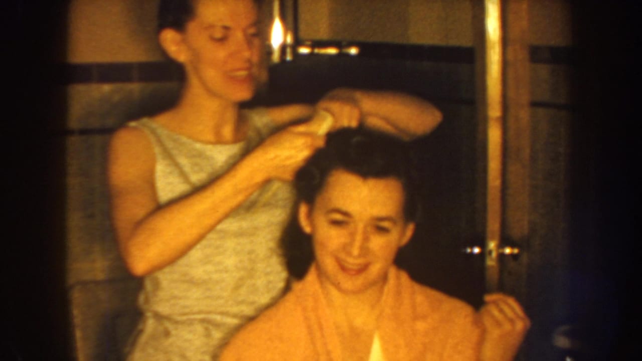 In 1959 Nebraska home-movie footage, a woman in a sleeveless dress brushes a seated woman’s dark hair in a bathroom.