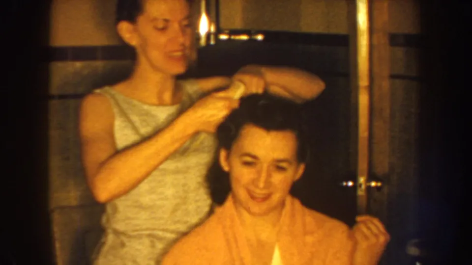 In 1959 Nebraska home-movie footage, a woman in a sleeveless dress brushes a seated woman’s dark hair in a bathroom.