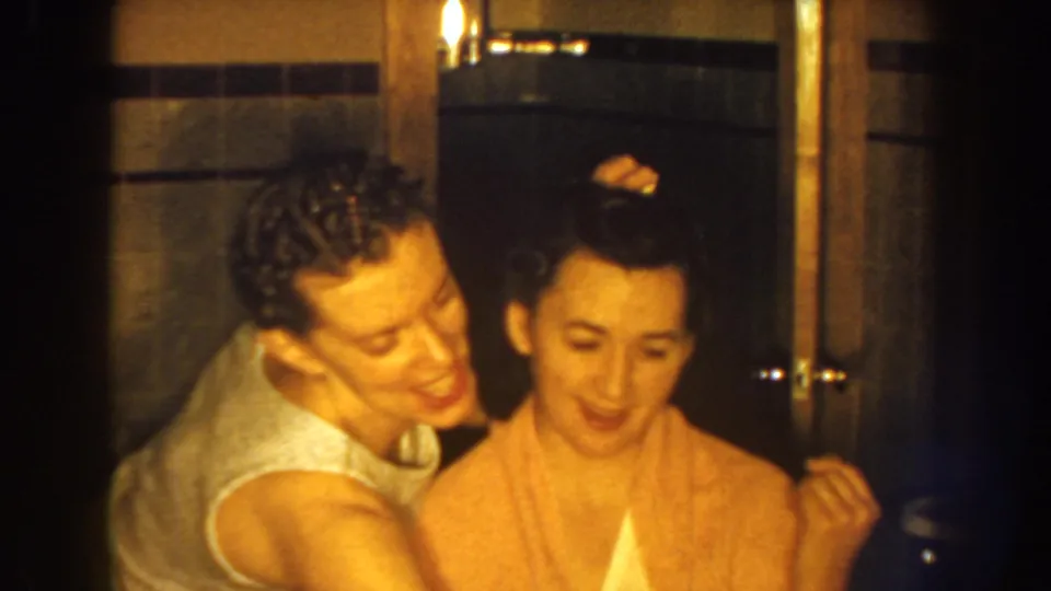 The stylist leans close to adjust the seated woman’s hair in a private bathroom, with tile and wood trim visible.