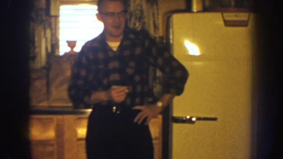 A man wearing glasses and a plaid shirt stands in a Nebraska kitchen near a rounded vintage refrigerator.