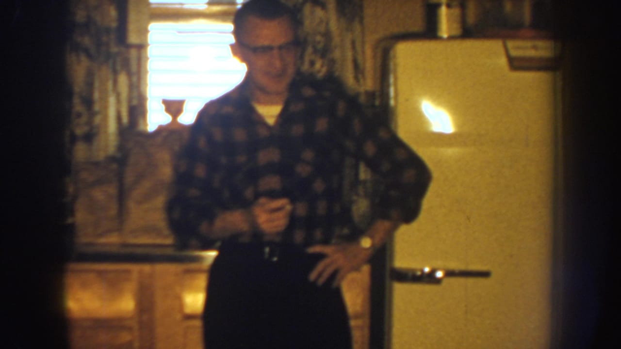 Adult man in glasses stands in a 1959 Nebraska kitchen beside a rounded white refrigerator, with cabinets and a bright window behind him.
