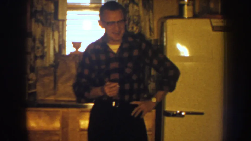 Adult man in glasses stands with one hand on his hip in a 1959 Nebraska kitchen, framed by wood cabinets, window blinds, and a white refrigerator.