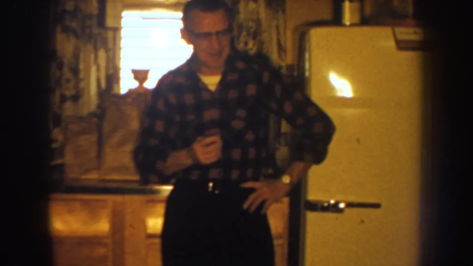 Adult man in a plaid shirt faces the camera near a white refrigerator in a warm, grainy Nebraska home-movie kitchen scene.