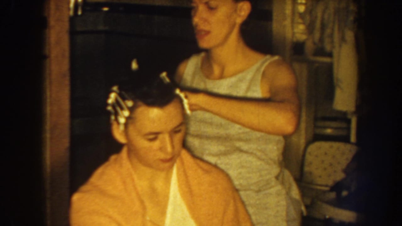 Nebraska 1959 home-movie frame of a woman setting hair rollers for a seated woman wrapped in a yellow towel.