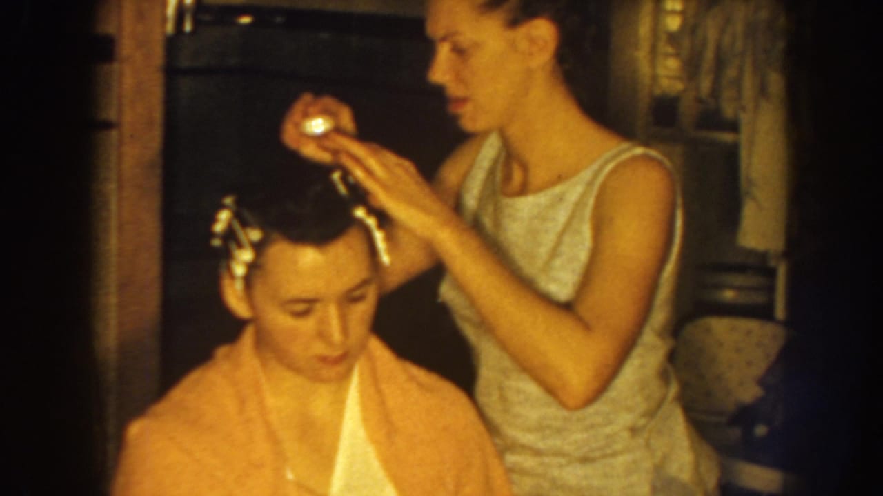 A standing woman adjusts a roller on the seated woman's hair.