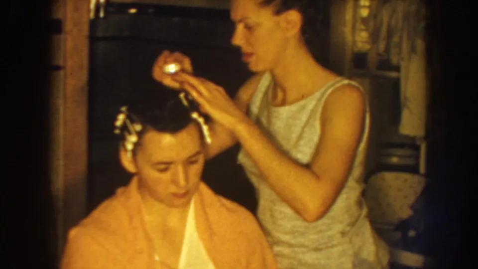 Nebraska 1959 frame showing a standing woman positioning a hair roller on another woman's head indoors.