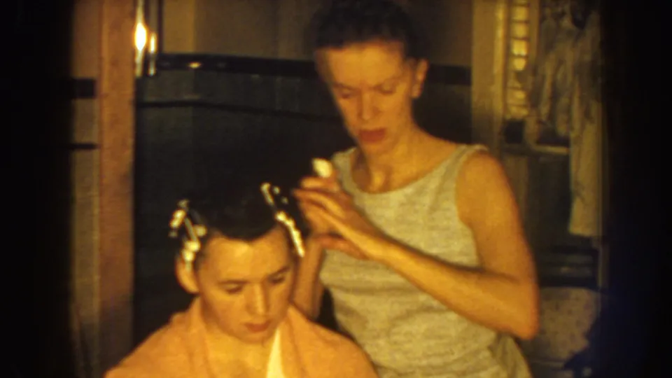 Nebraska 1959 home-movie frame with one woman concentrating as she sets rollers in another woman's hair.