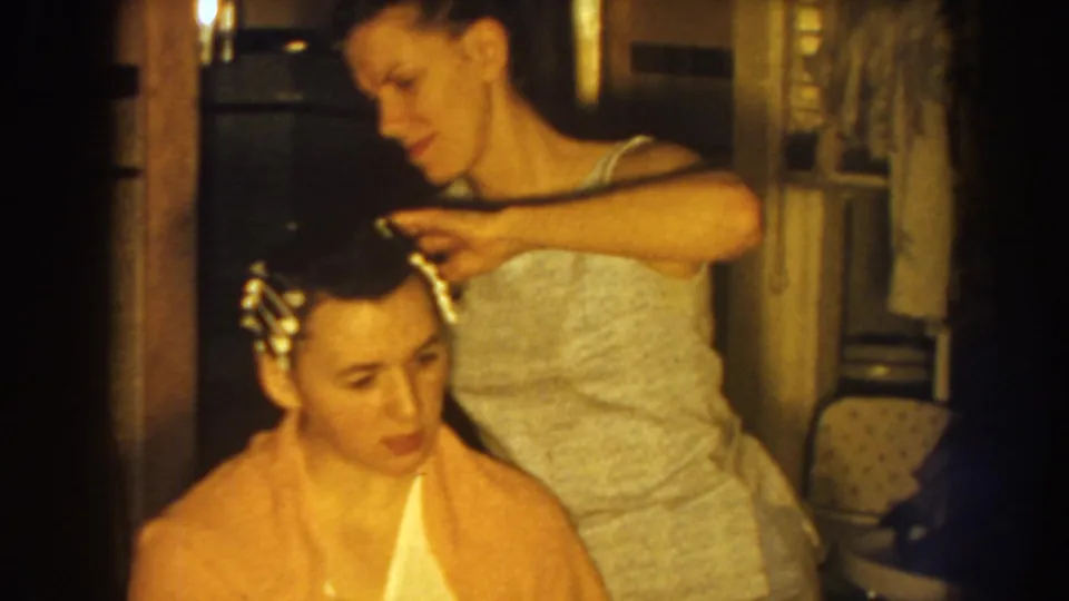 A woman in a light sleeveless top carefully sets curlers in another woman's hair inside a Nebraska home interior.