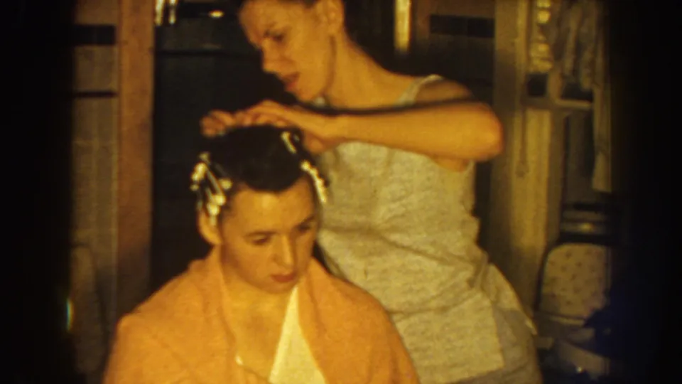 The seated woman, wrapped in a pink towel, looks down as another woman continues placing curlers in her hair.