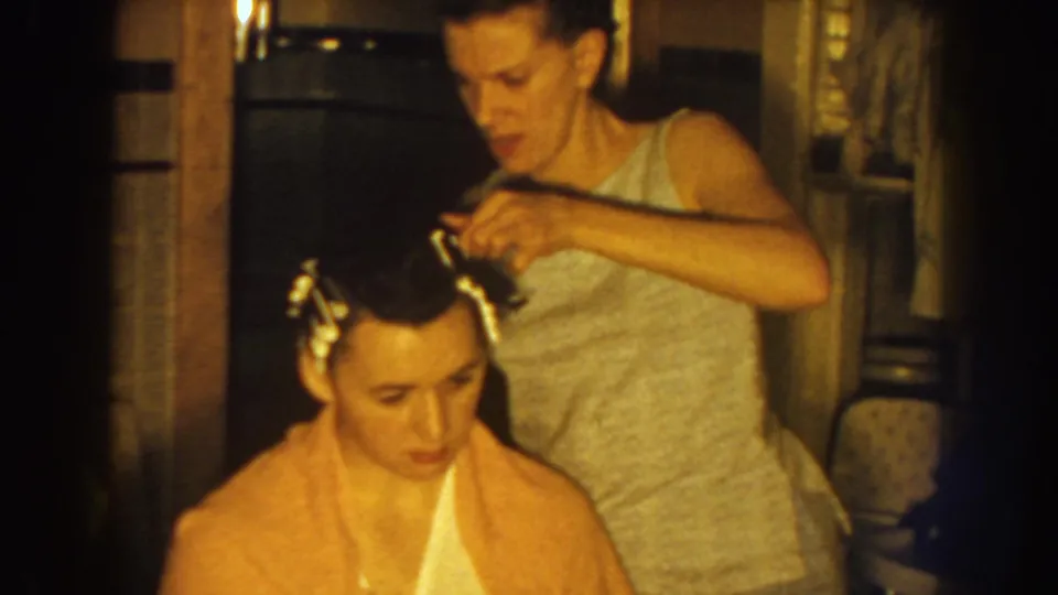 Near the end of the clip, the standing woman leans in to finish adjusting curlers on the seated woman's hair.