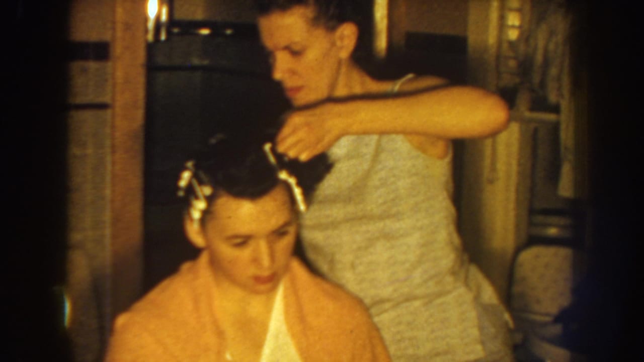In metadata-supported 1959 Nebraska home-movie footage, a woman in a light top places curlers in the hair of a seated woman wrapped in a peach towel.