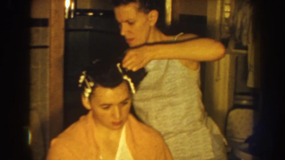 In metadata-supported 1959 Nebraska home-movie footage, a woman in a light top places curlers in the hair of a seated woman wrapped in a peach towel.