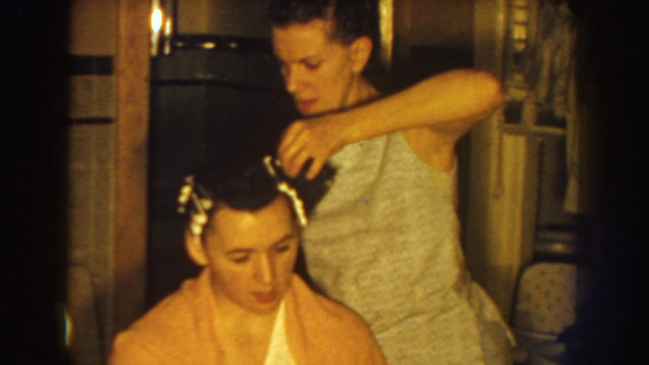 Standing woman adjusts curlers as the seated woman looks downward in an interior room.
