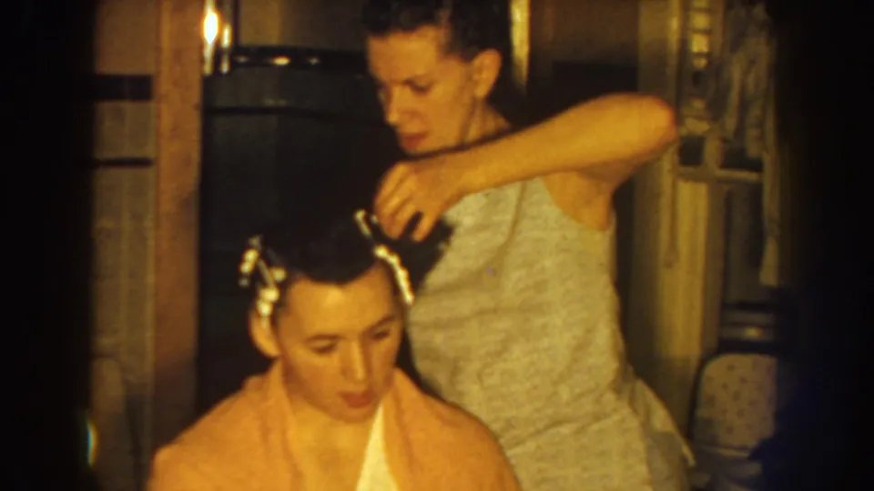 A woman carefully adjusts hair curlers on a seated woman wrapped in a peach towel, filmed indoors in metadata-supported 1959 Nebraska footage.