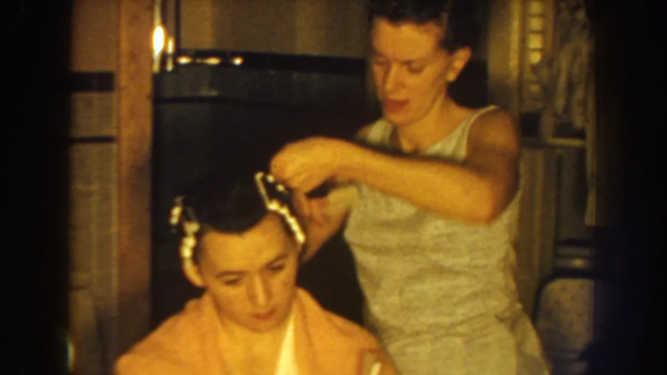 The standing woman reaches over the seated woman's head, continuing to set curlers in a private indoor grooming scene from metadata-supported 1959 Nebraska footage.
