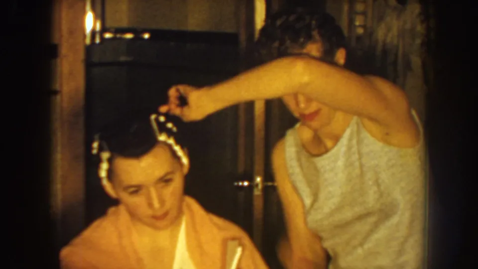 A woman lifts hair near the curlers as the seated woman tilts her head, continuing the home grooming routine in metadata-supported 1959 Nebraska footage.