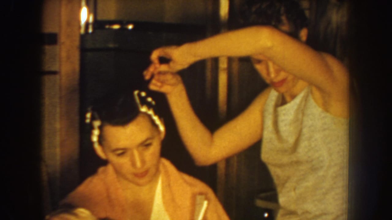In 1959 Nebraska home-movie footage, a woman reaches over a seated woman with hair rollers in a warm indoor room.