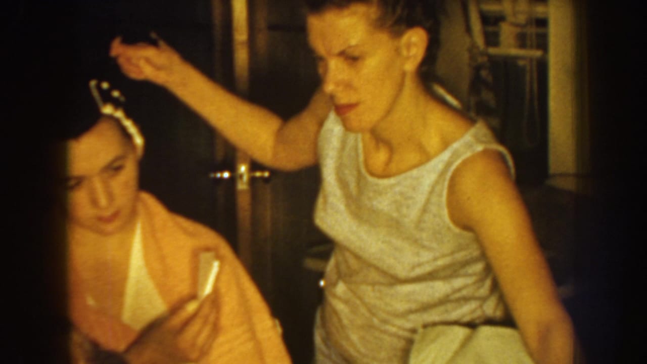 Seated woman with rollers holds an object while another woman styles her hair.