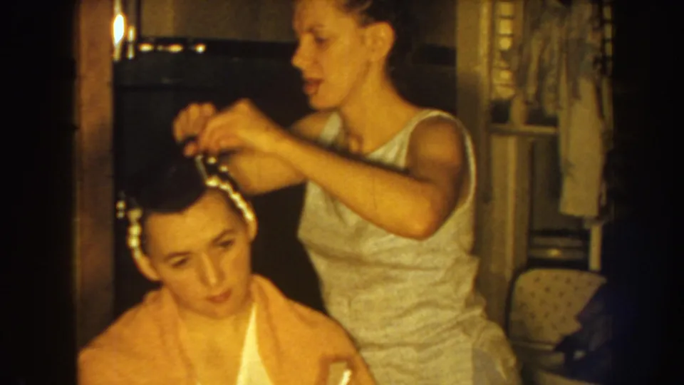 In the final frame, the standing woman adjusts hair rollers while the seated woman looks downward in a private Nebraska interior.