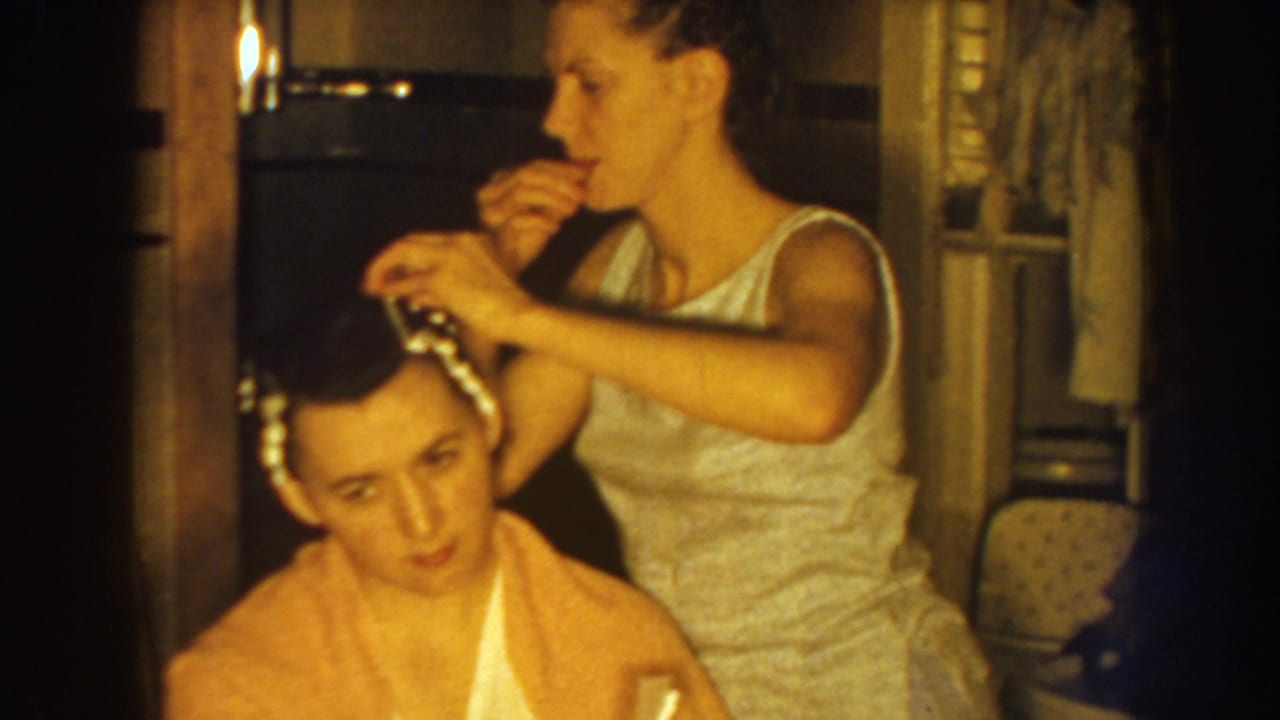 In 1959 Nebraska home-movie footage, a woman in a sleeveless dress works on a seated woman's hair in a warm private interior.
