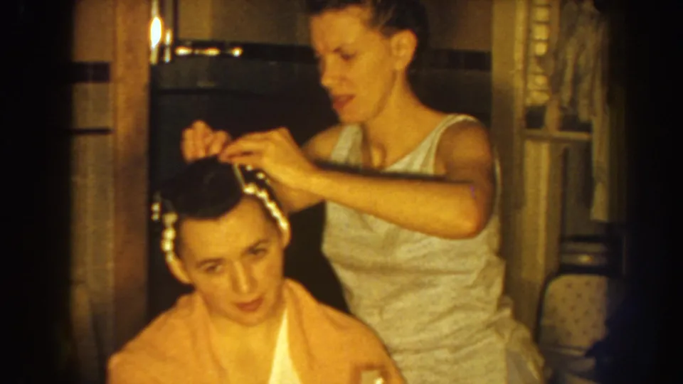 A woman carefully works near the seated woman's hairline, with clips visible against the dark hair in the 1959 home footage.