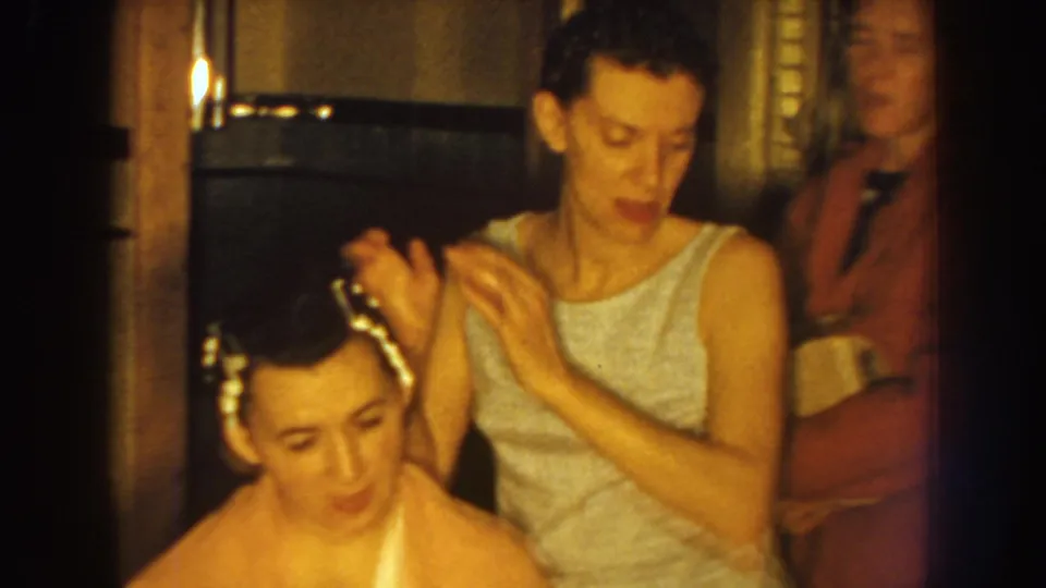 The woman pauses after arranging the hair, while another adult figure appears partly at the edge of the private interior frame.