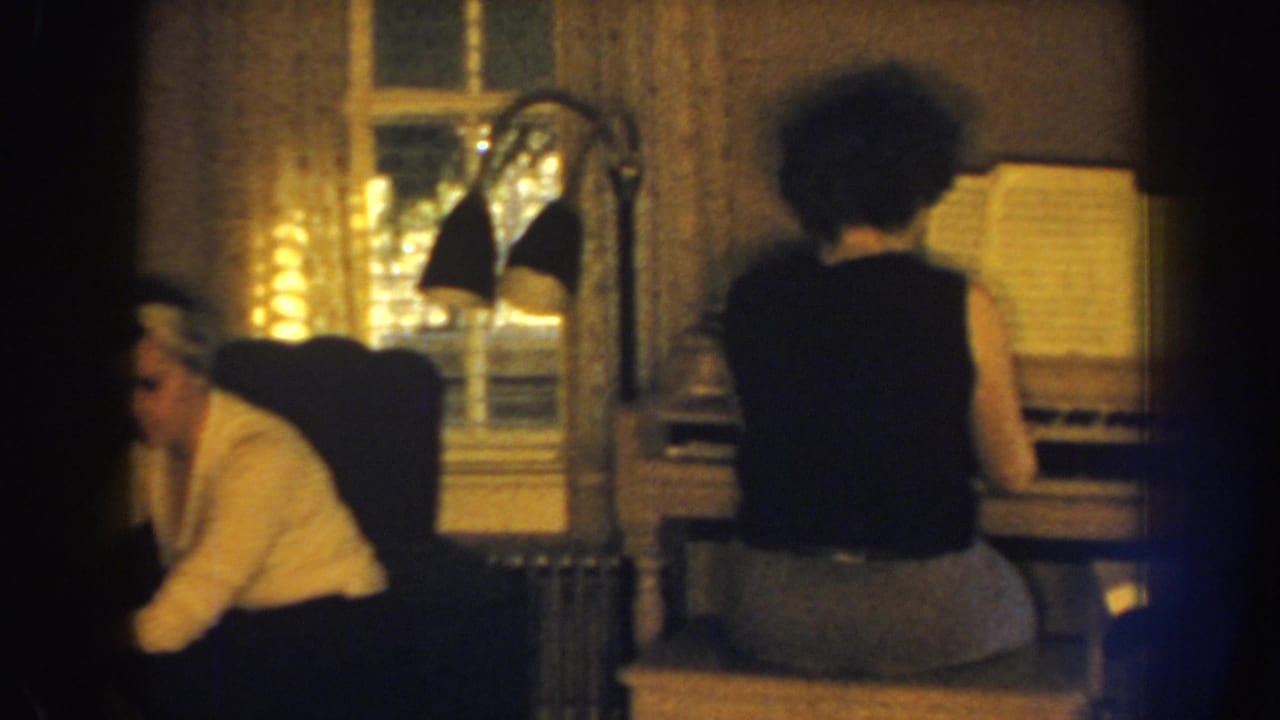 1959 Nebraska home-movie frame showing adults inside a private living room, including a person at an organ or piano and a seated woman nearby.