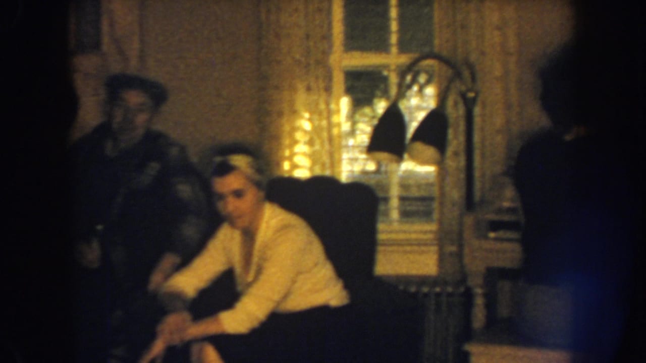 A woman sits in a chair while a man in a cap stands nearby in a warmly lit room.