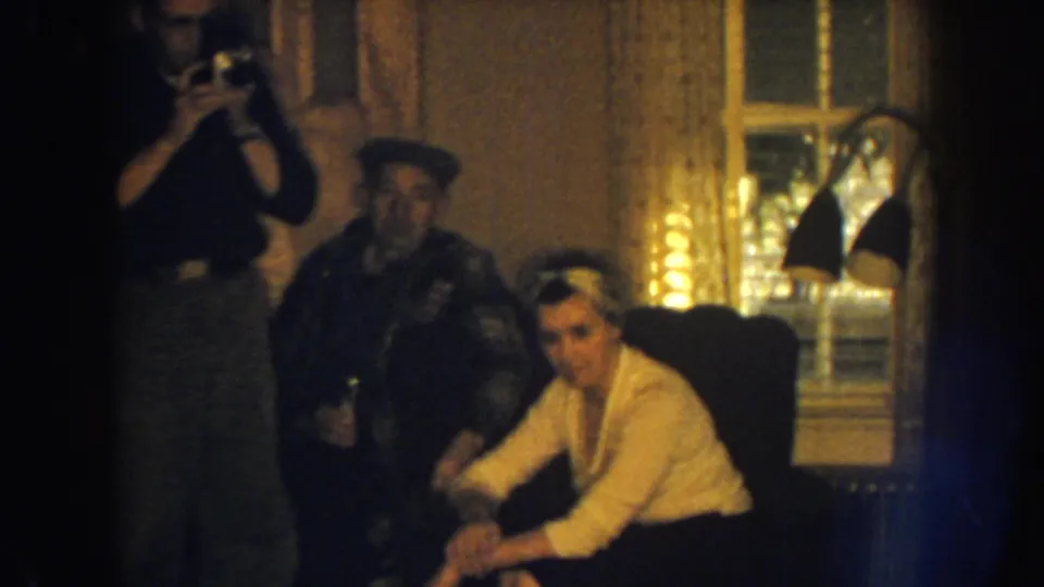 1959 Nebraska home-movie frame showing a man with a still camera photographing adults gathered in a warmly lit private living room.