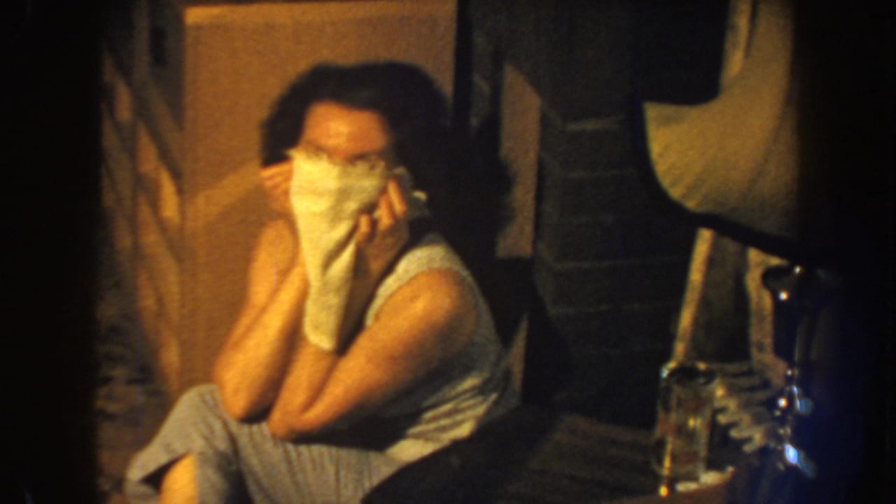 In 1959 Nebraska home-movie footage, a seated woman in a sleeveless top and striped pants covers her face with a white cloth indoors.