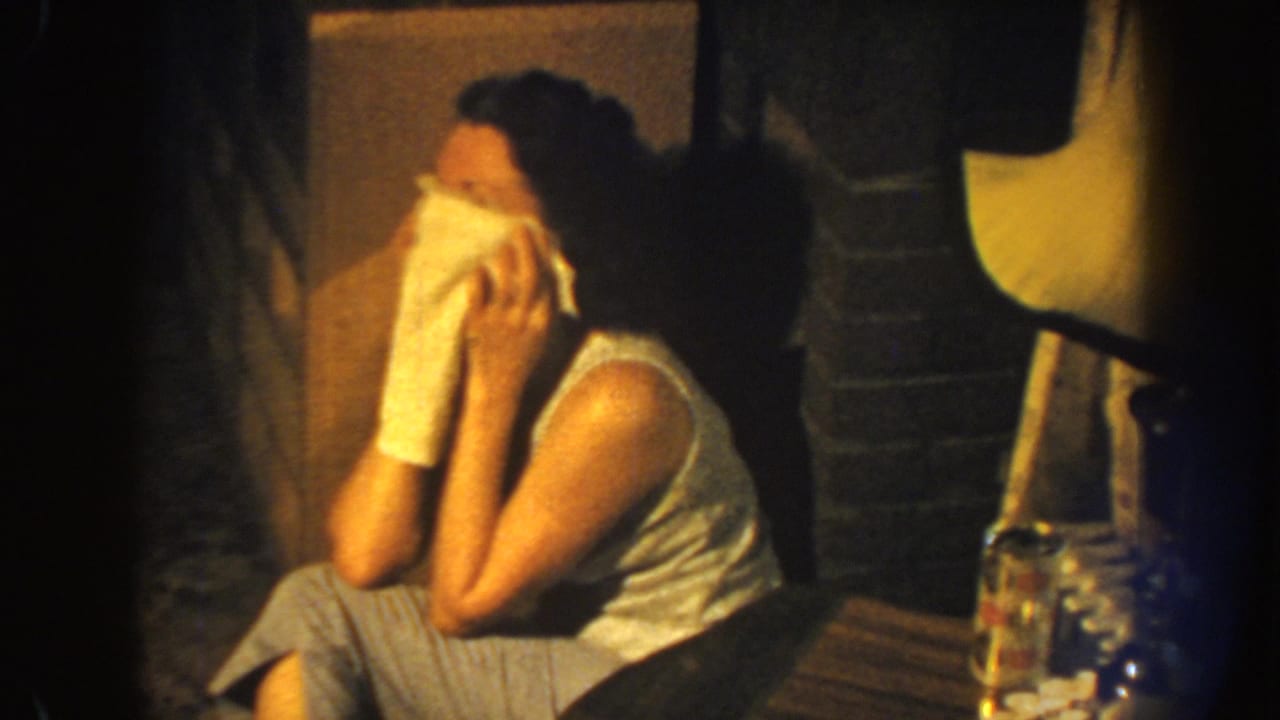 The woman remains seated in profile, holding a white cloth to her face beside a small table and glass container in a dim room.
