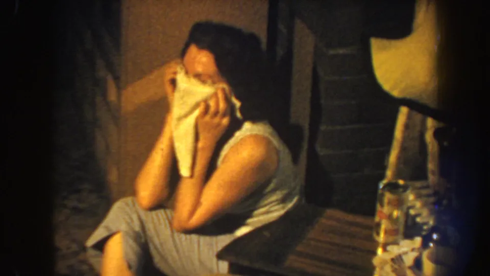 The seated woman leans forward with the cloth still raised, her posture framed by warm 8mm grain and a shadowed interior.