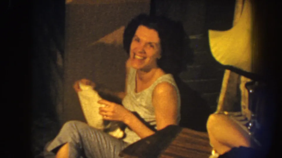 The woman lowers the white cloth and smiles toward the camera, revealing a candid domestic moment in 1959 Nebraska footage.