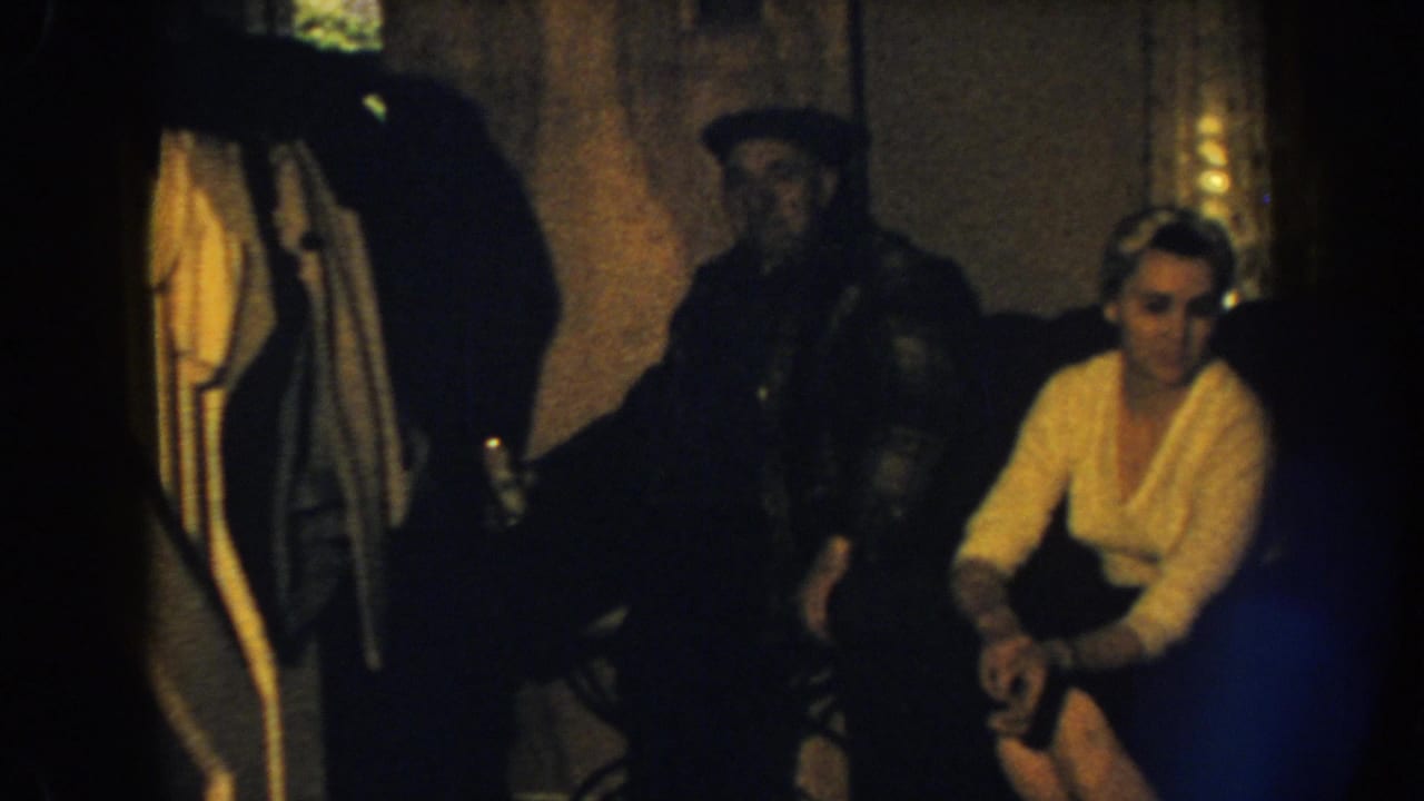 1959 Nebraska home-movie frame showing adults on a sofa in a dim interior, with one partial figure standing near the left edge.