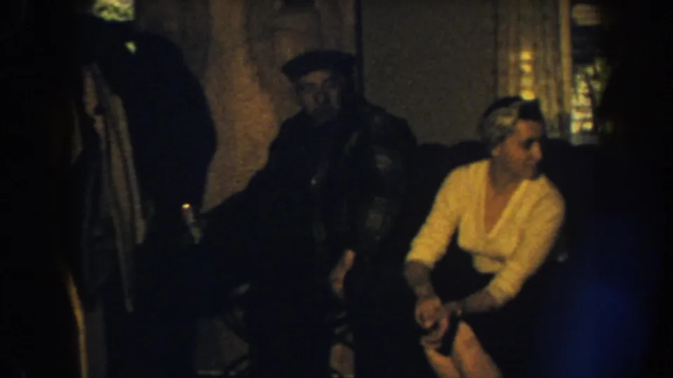 1959 Nebraska interior frame with a woman in a light sweater seated beside a man in dark clothing and a cap on a sofa.