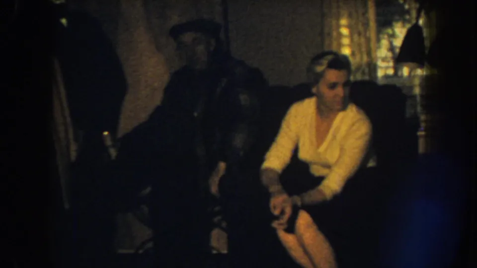 1959 Nebraska home-movie frame with a woman in a light sweater seated on a sofa and a shadowed man beside her.