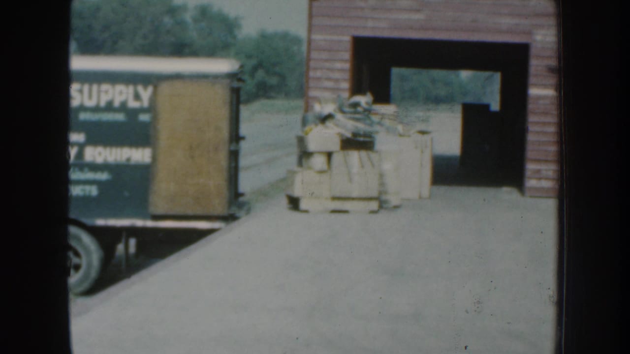 1959 Nebraska 8mm frame showing a commercial loading dock, stacked materials, a partial supply truck, and a red-sided building exterior.