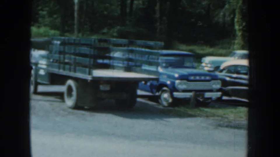 1959 Nebraska 8mm frame showing a blue pickup and late-1950s cars parked in a lot bordered by trees.