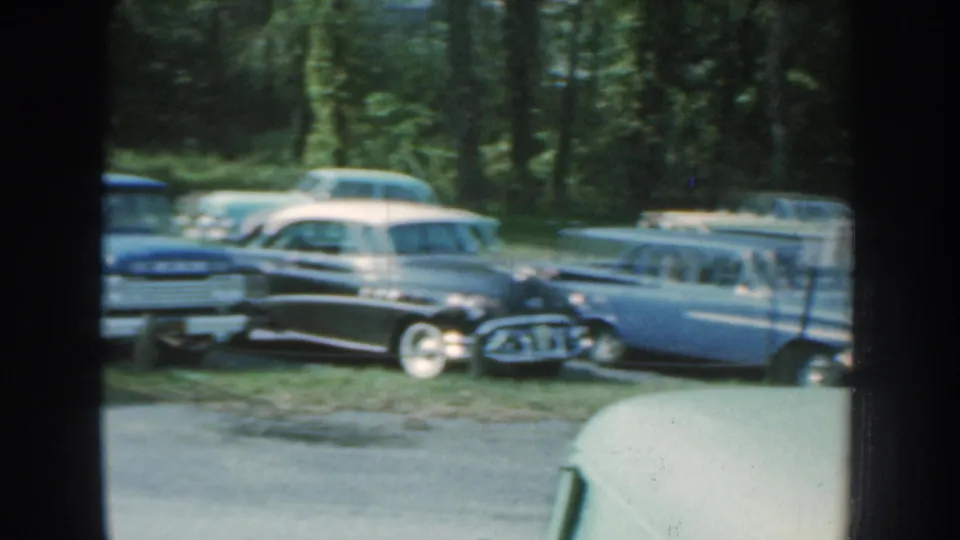 Nebraska 1959 frame of several late-1950s cars parked near trees, with part of a light-colored vehicle in the foreground.